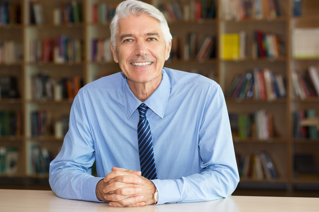 photo of a middle aged man in a shirt and tie smiling at the camera while seated at a desk