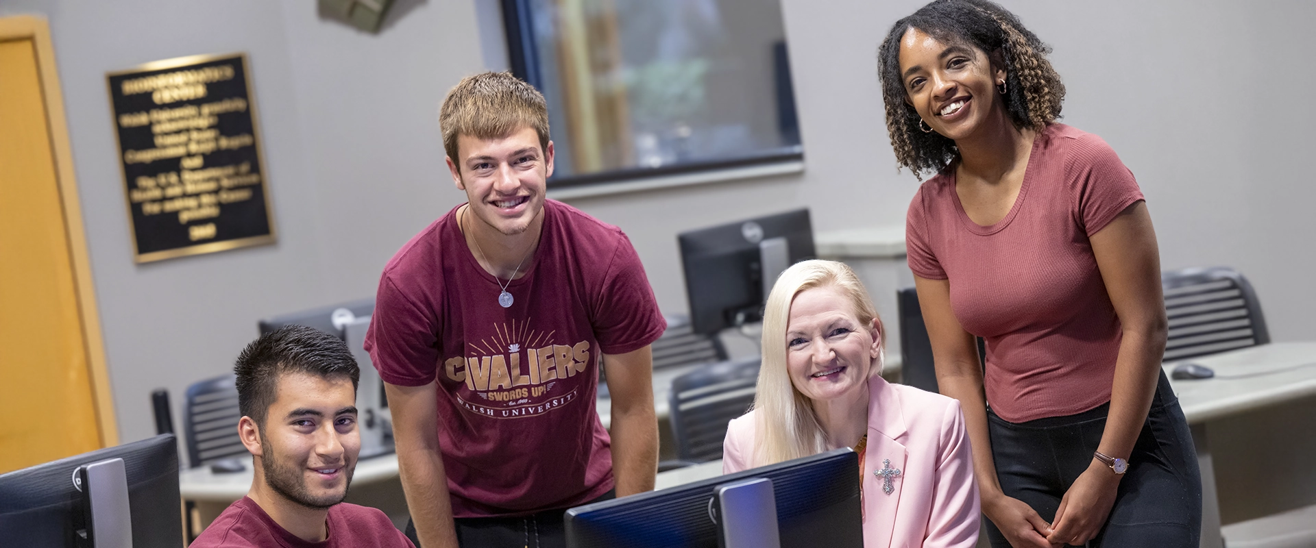 Three students and a professor positioned behind a computer screen while posed and smiling at the viewer