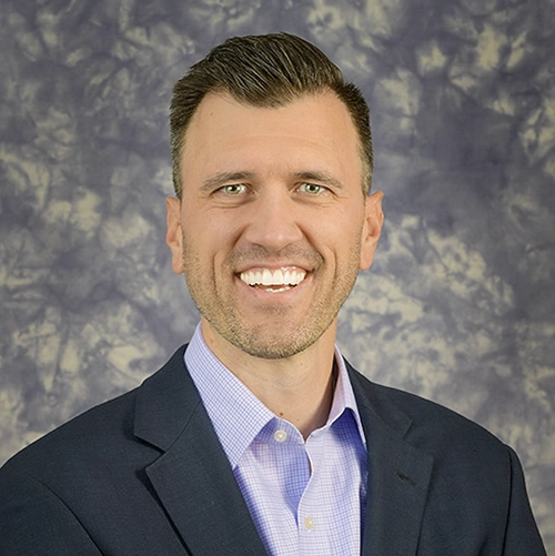 Photo of Adam Lesch, a professional portrait of a smiling man in a suit with a collared shirt against a textured background.