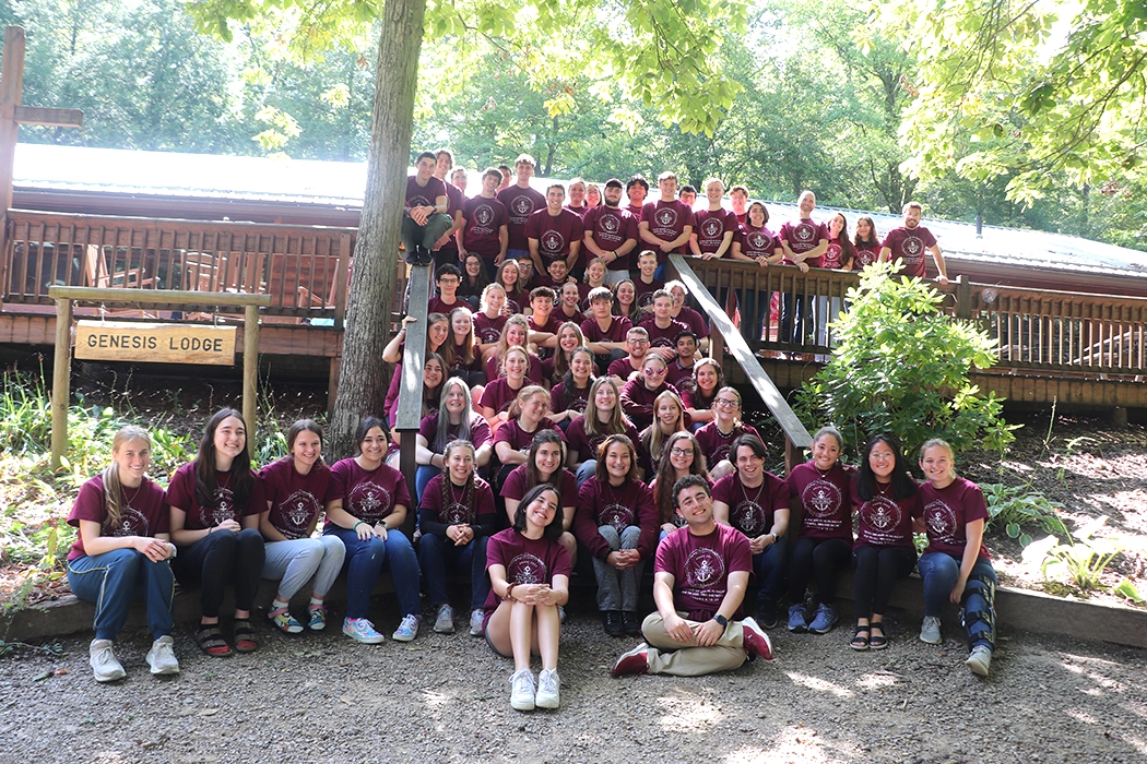 Group photo of a large gathering of participants wearing matching maroon t-shirts, seated and standing in front of the Genesis Lodge amidst a wooded area.