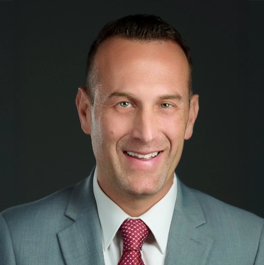 Portrait of David Shaffer, a smiling professional in a gray suit with a red tie against a dark background.