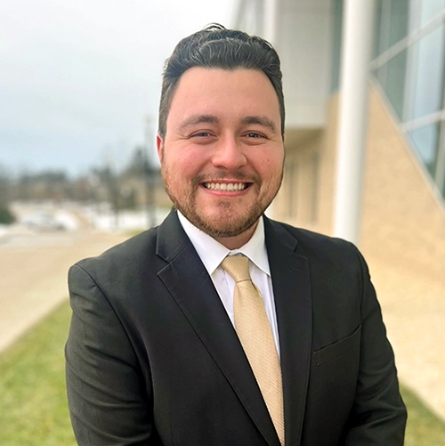 Photo of Gannon Fridley, a smiling young man in a black suit and beige tie stands outdoors, showcasing a professional appearance.