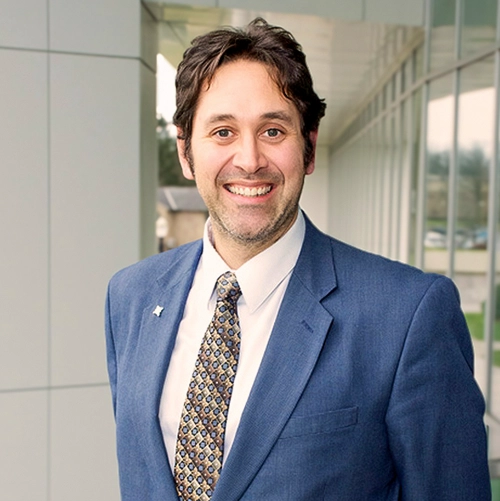 Professional headshot of Miguel Chavez, a man in a suit, smiling confidently against a modern architectural backdrop.