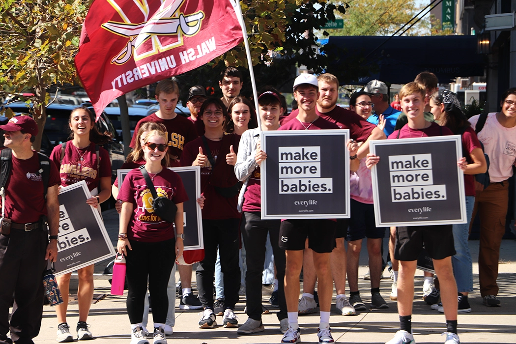 A group of university students in maroon apparel holding signs that say "make more babies" and a large flag, participating in a lively demonstration on a city street.