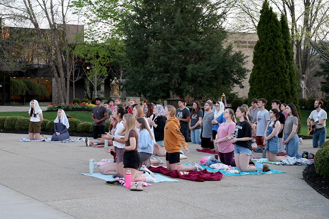 A group of people gathered outdoors for a prayer session, some kneeling and others standing, in a serene setting surrounded by trees.