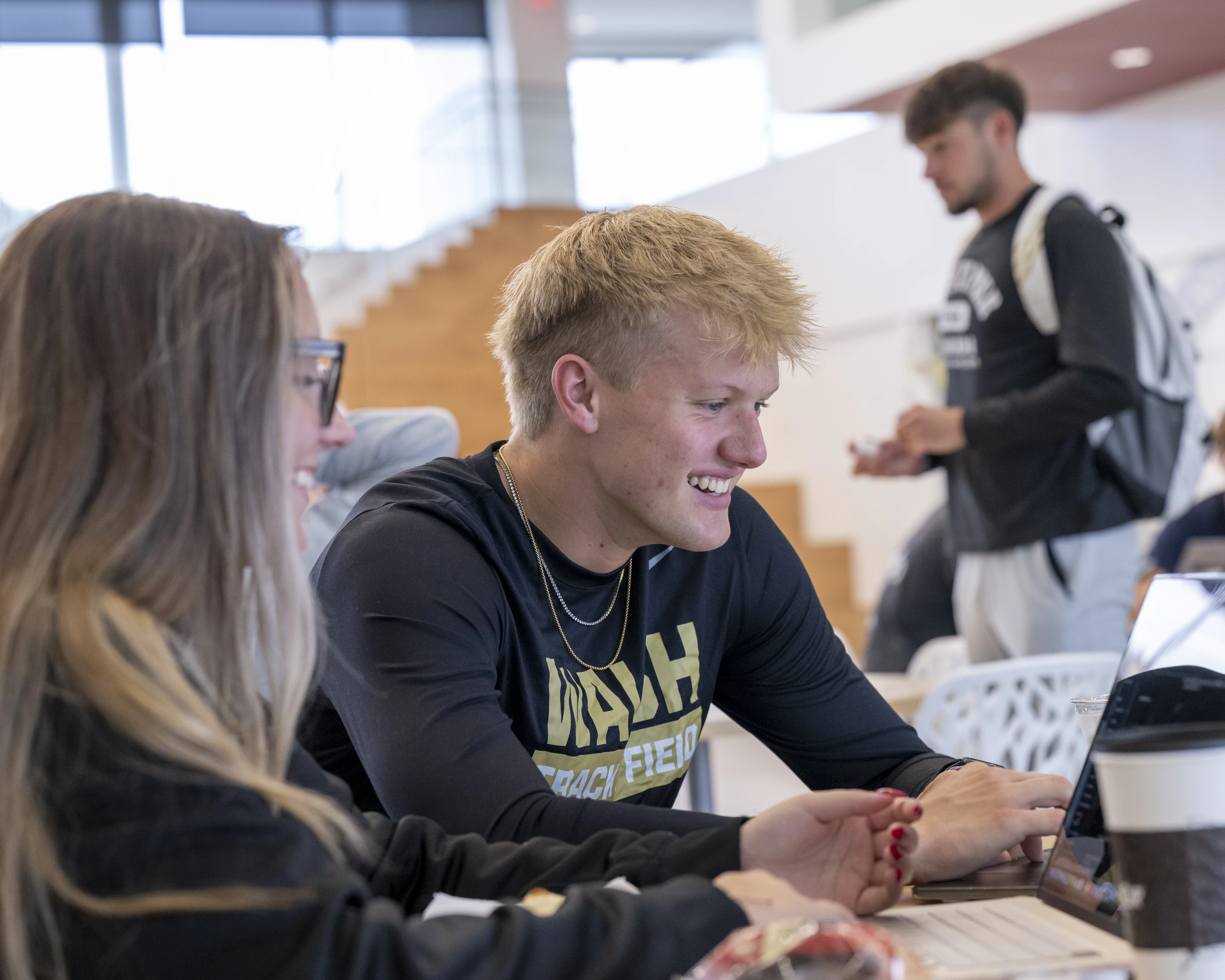 Two students seated in a brightly lit area while working on a computer and having a conversation