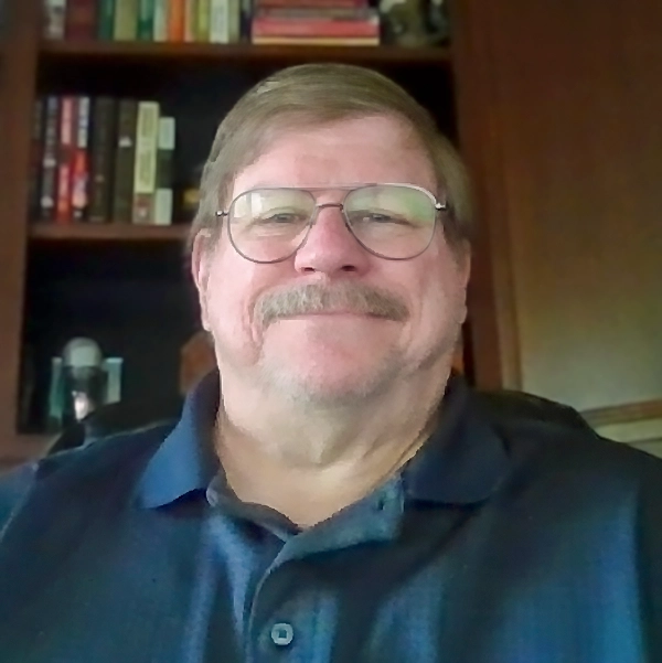 Photo of Thomas Herrick, a smiling man with glasses and a beard, sitting in front of a bookshelf filled with books.