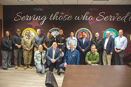 A group photo of military and civilian personnel in front of a wall that reads "Serving those who Served."
