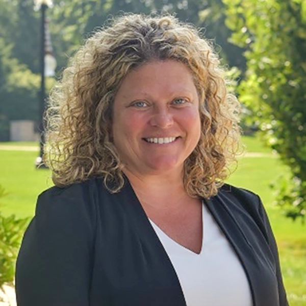 Portrait of a smiling woman with curly hair, dressed in a black blazer against a green outdoor background.
