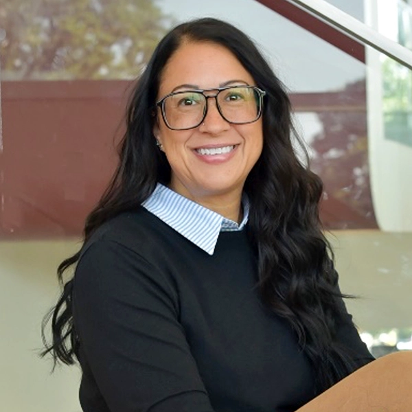 A smiling woman with long, wavy hair and glasses, seated in a modern setting, wearing a black sweater with a collared shirt underneath.