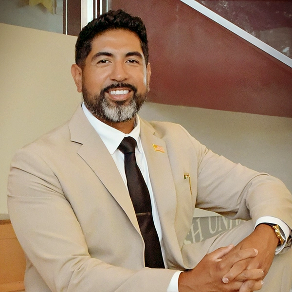 Portrait of a smiling man in a beige suit with a tie, sitting confidently with one arm resting on his knee.