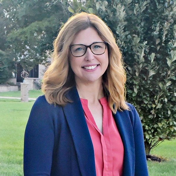 A professional headshot of a smiling woman with glasses, wearing a navy blazer over a coral blouse, set against a green landscape.