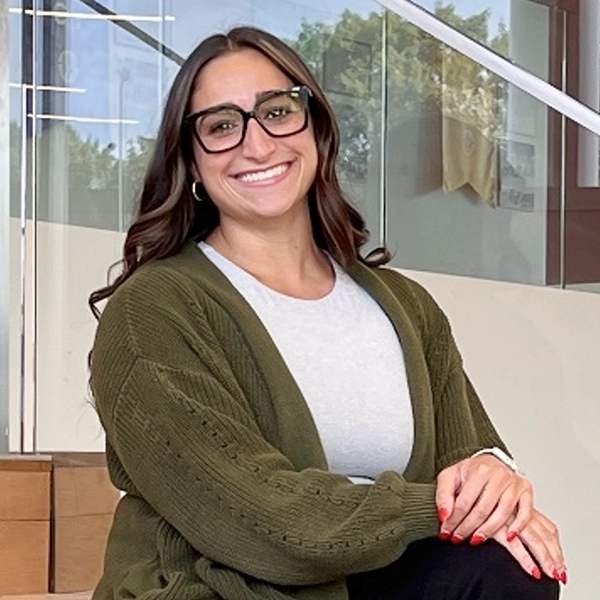 Smiling woman with glasses and long hair, seated in a modern indoor setting, wearing a green cardigan over a gray shirt.