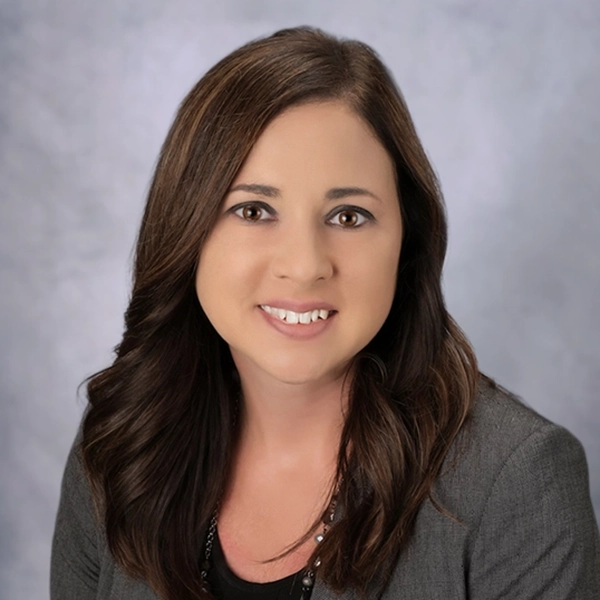 Professional headshot of a woman with long brown hair, wearing a grey blazer and black shirt.