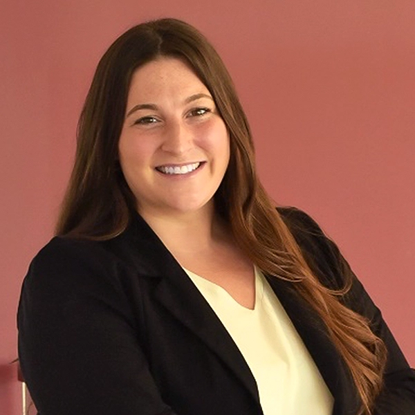 Professional portrait of a smiling woman in a blazer against a solid colored background.