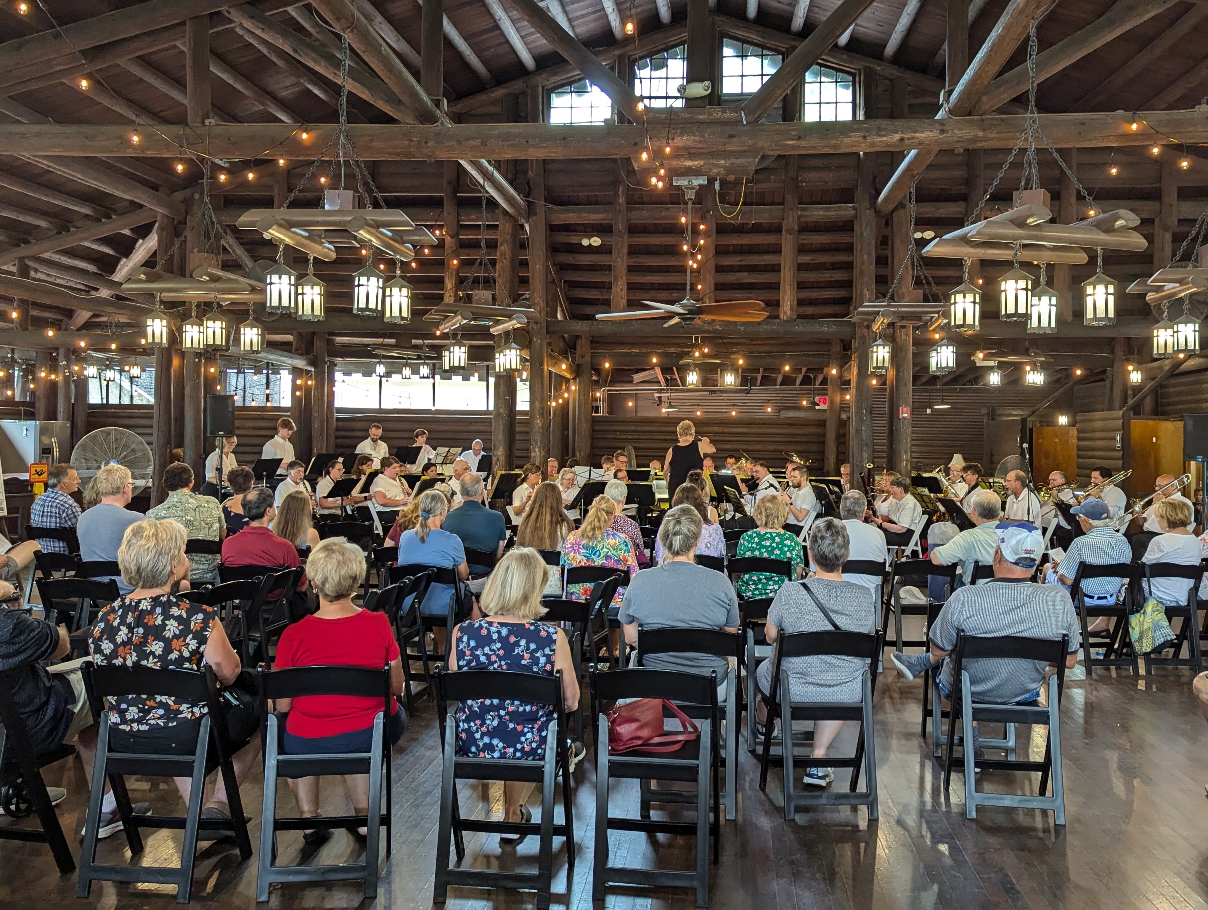 Audience seated on folding chairs watches a concert band perform inside a rustic wooden hall with hanging lanterns and string lights.