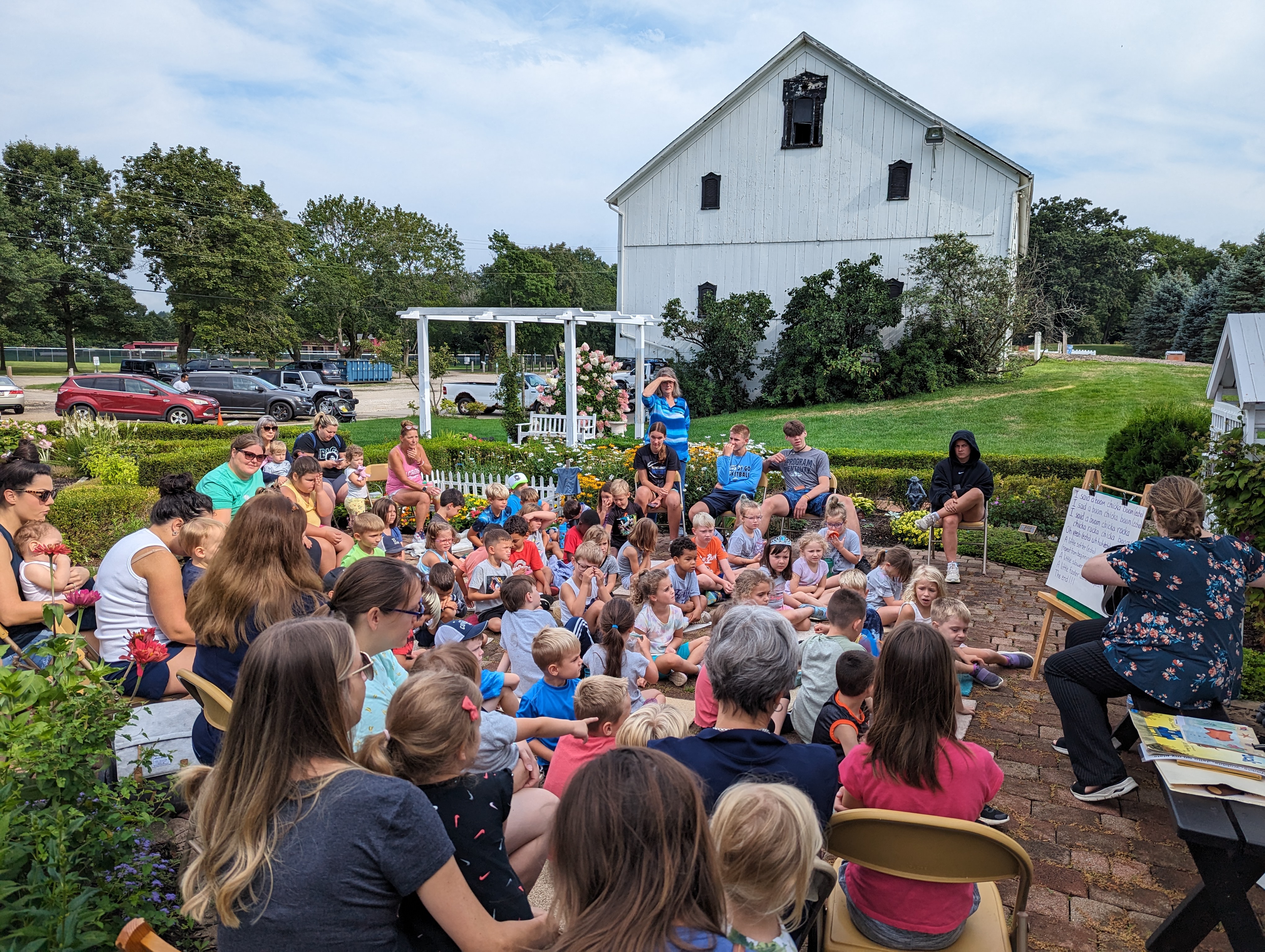 Group of children and adults seated outdoors for a storytime or presentation in a garden courtyard, with a large white barn in the background.