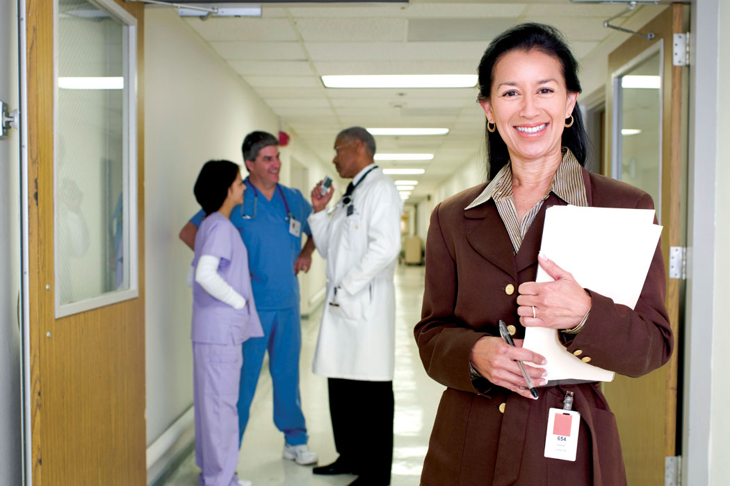 Hospital administrator standing in a hallway smiling at the camera while a doctor and two nurses consult with each other in the background
