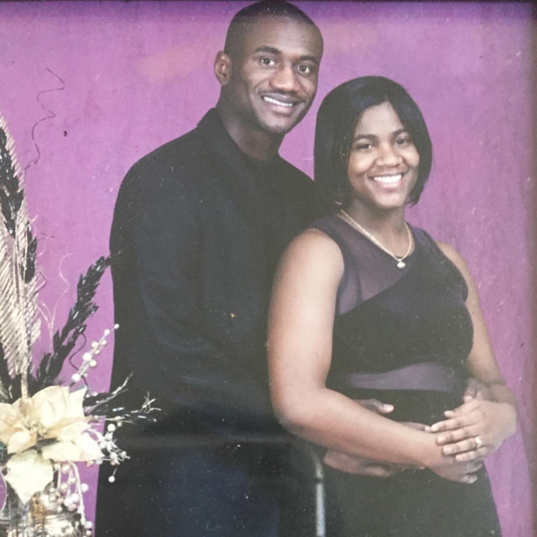 A couple posing together in formal attire, smiling against a purple backdrop. The woman is wearing a black dress, and the man is in a black suit.