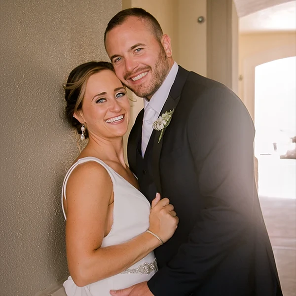 A happy couple smiles together, posing for a romantic portrait. The woman is wearing a white dress, and the man is in a formal suit with a boutonnière.