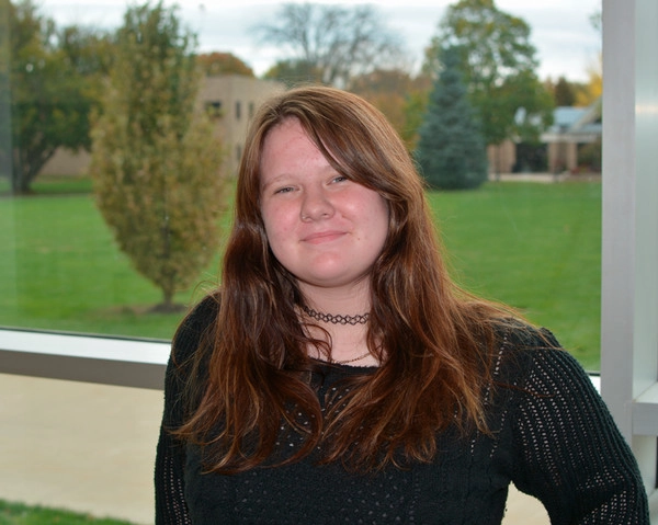 A smiling student with long brown hair standing near a large window, showcasing a grassy outdoor area in the background.