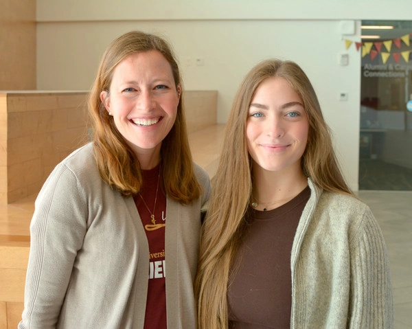Two women smiling together in a modern indoor space. One is wearing a university-themed shirt while the other has long hair and a cozy sweater.