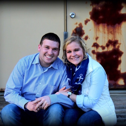 A smiling couple poses together, dressed casually against a backdrop with a rusty texture.