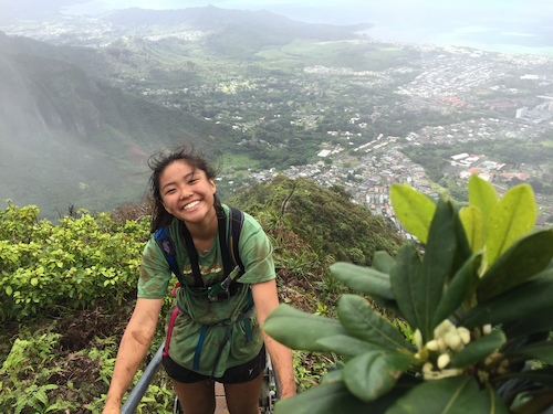 A smiling hiker climbs a scenic trail overlooking a lush valley and coastline.